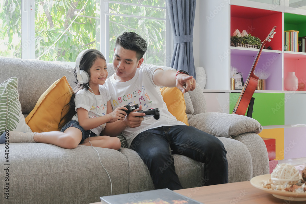 Two asian young father and adorable little girl daughter family enjoy playing game console together with happy smiling face at sofa living room on holiday weekend. Selective focus. Two asian young father and adorable little girl daughter family enjoy playing game console together with happy smiling face at sofa living room on holiday weekend. Selective focus.