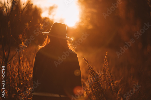 A young girl in a hat stands against the background of the sunset in the field and admires the light sun