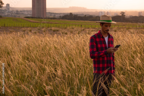Farmer with hat and mobile tablet analyzing the plantation on sunset. Buildings and city blurred background. Space for text.