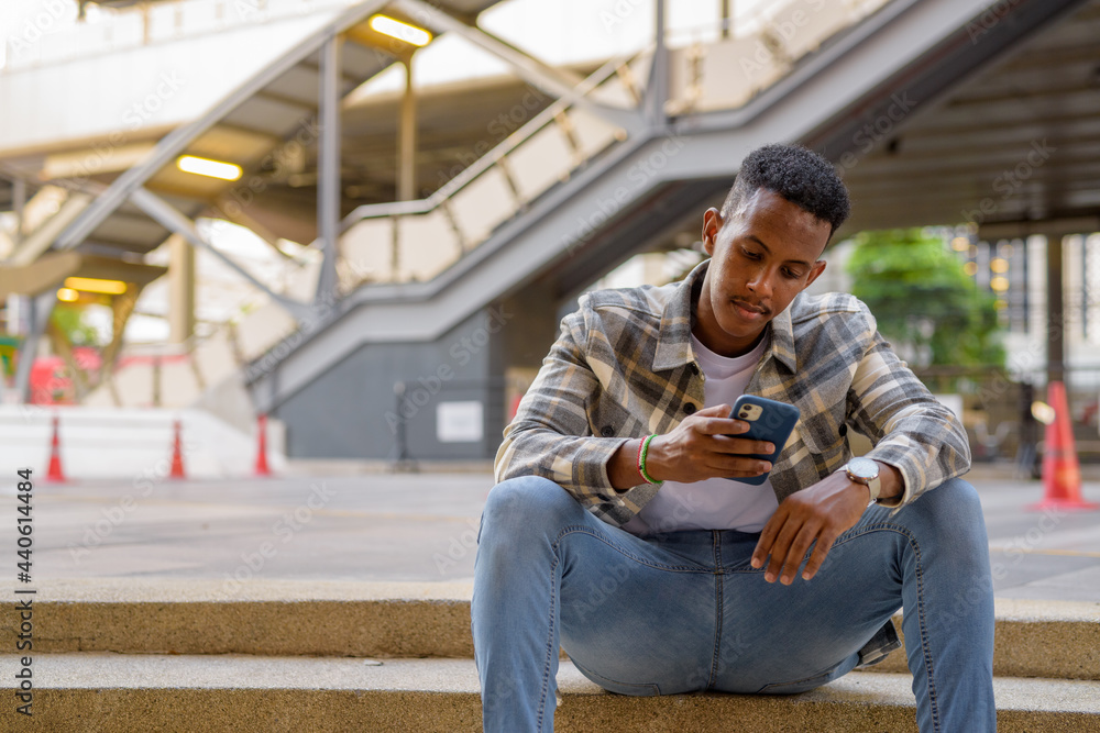Portrait of African black man sitting outdoors in city during summer using mobile phone