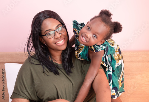 Obraz na plátně Head shot portrait of happy African American mother cuddles a lovely little daughter sitting on the sofa and looking at camera
