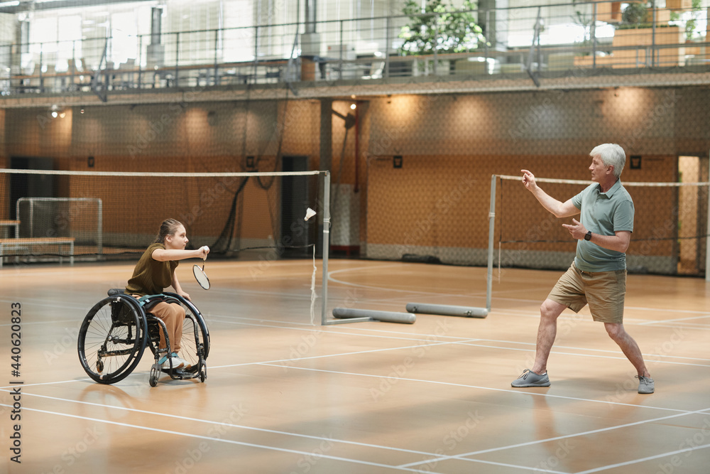 Full length portrait of senior coach training young woman in wheelchair during badminton practice in sports court, copy space