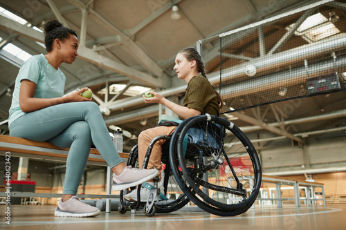 Full length portrait of young woman in wheelchair sharing lunch with friend at indoor sports court