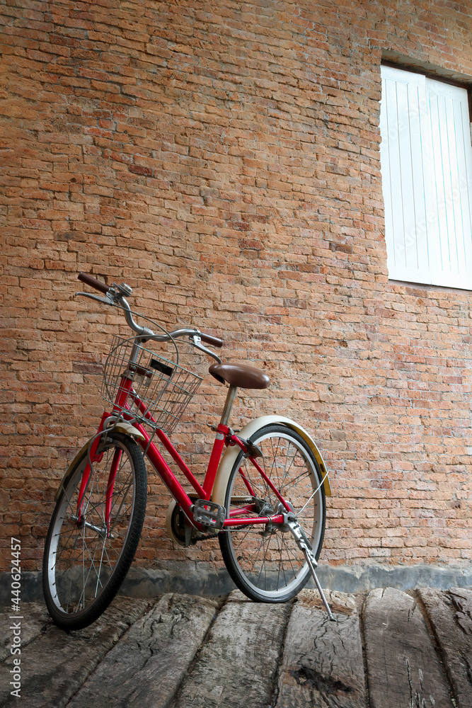 Red vintage classic bicycle with white basket, brown saddle seat and wheels. Bike park in front of brick wall with white wooden window on grunge aged wood floor.