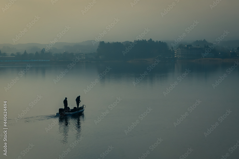 Silhouette of two male fisherman sailing boat in the lake for fishing with landscape of wild forest in the background. Tree and man shadow reflect on water. Thick fog in morning with blue color tone.