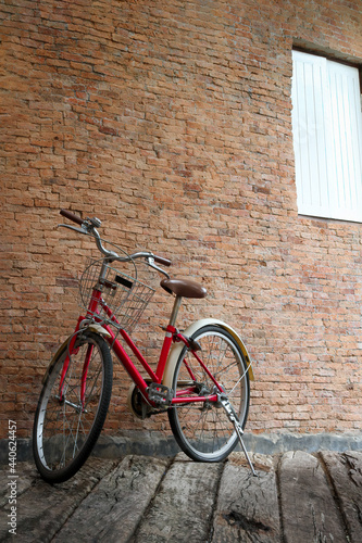Red vintage classic bicycle with white basket, brown saddle seat and wheels. Bike park in front of brick wall with white wooden window on grunge aged wood floor.