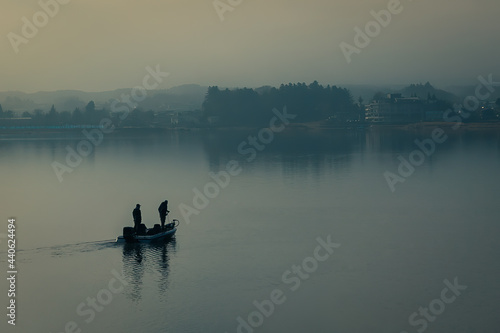 Silhouette of two male fisherman sailing boat in the lake for fishing with landscape of wild forest in the background. Tree and man shadow reflect on water. Thick fog in morning with blue color tone.