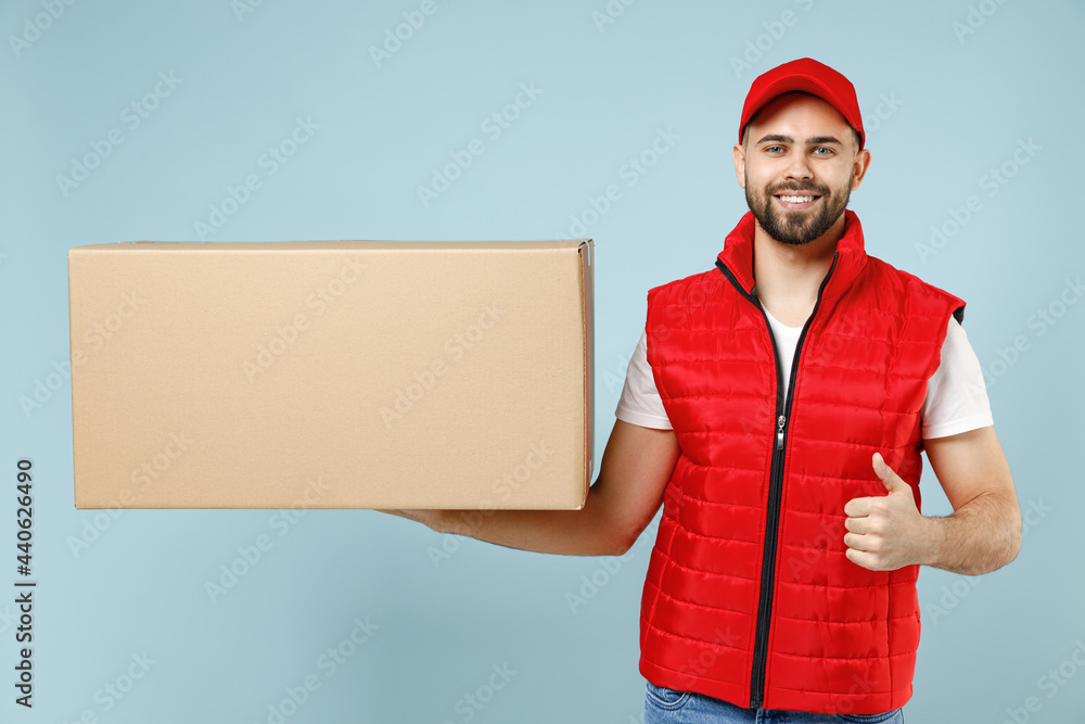 Professional delivery guy employee man in red cap white T-shirt vest uniform workwear work as dealer courier hold blank cardboard box isolated on pastel blue color background studio. Service concept. Professional delivery guy employee man in red cap white T-shirt vest uniform workwear work as dealer courier hold blank cardboard box isolated on pastel blue color background studio. Service concept.