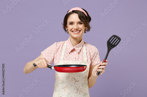 Young smiling happy fun cheerful housewife housekeeper chef cook baker woman in pink apron hold show red frying pan spatula isolated on pastel violet background studio Cooking food process concept photo