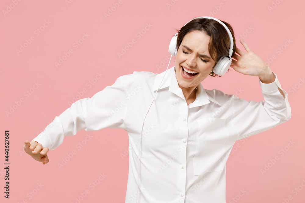 Young joyful successful employee business woman corporate lawyer 20s wear classic formal white shirt headphones work in office dance listen to music isolated on pastel pink background studio portrait. Young joyful successful employee business woman corporate lawyer 20s wear classic formal white shirt headphones work in office dance listen to music isolated on pastel pink background studio portrait.