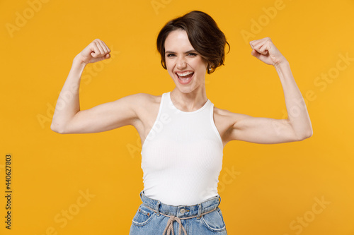Young strong sporty fitness woman 20s with bob haircut wearing white tank top showing biceps muscles on hand demonstrating strength power isolated on yellow color background. People lifestyle concept.