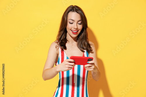 Young woman wear striped red blue swimsuit using play racing on mobile cell phone hold gadget video games isolated on vivid yellow color background studio Summer hotel pool sea rest sun tan concept