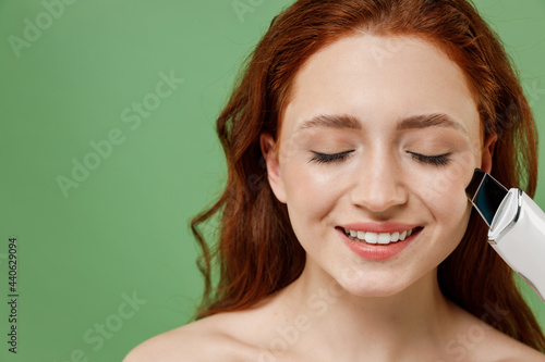 Beautiful smiling half naked topless redhead hair woman 20s with nude make up hold ultrasonic face scrubber isolated on pastel green color background Skin care healthcare cosmetic procedures concept.