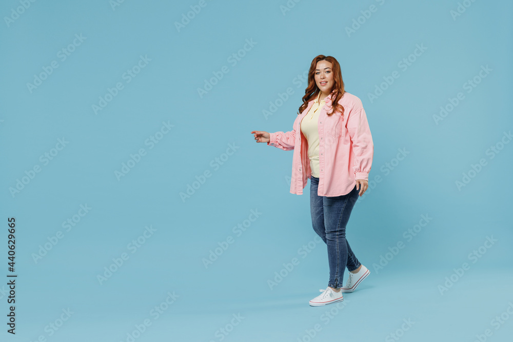 Full length side view young fun smiling redhead chubby overweight woman 30s wearing in pink shirt jeans casual clothes walking going looking camera isolated on pastel blue background studio portrait Full length side view young fun smiling redhead chubby overweight woman 30s wearing in pink shirt jeans casual clothes walking going looking camera isolated on pastel blue background studio portrait