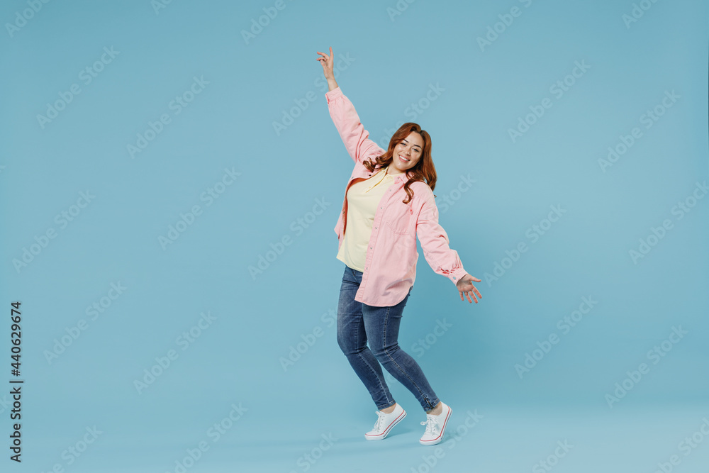 Full length young fun joyful smiling redhead chubby overweight woman 30s wearing in pink shirt jeans leaning back stand on toes with oustretched hands hands dance isolated on pastel blue background. Full length young fun joyful smiling redhead chubby overweight woman 30s wearing in pink shirt jeans leaning back stand on toes with oustretched hands hands dance isolated on pastel blue background.