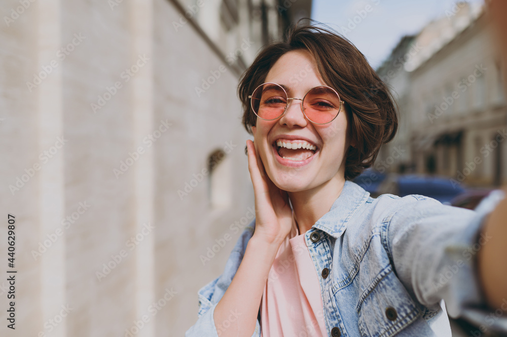 Close up young happy fun excited tourist caucasian woman 20s wear jeans clothes eyeglasses doing selfie shot on mobile phone on blur building background. People urban youth travel lifestyle concept
