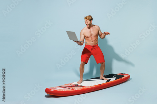 Canvas Print Full length young freelancer man 20s wear red shorts swimsuit stand on sup board point finger laptop pc computer surfing isolated on pastel blue background Summer vacation sea rest sun tan concept