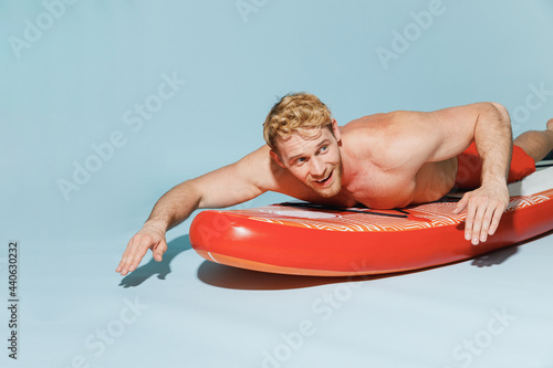 Canvas Print Young handsome sexy strong sporty blond man 20s in red shorts swimsuit lying on sup board paddle surfing isolated on pastel blue background studio portrait