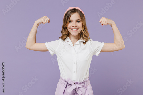 Little strong sporty fitness kid girl 12-13 year old in white shirt showing biceps muscles on hand demonstrate strength power isolated on purple background children studio Childhood lifestyle concept