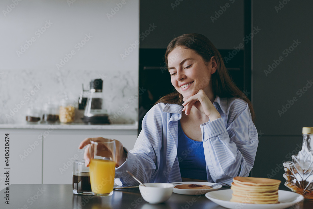 Young happy housewife woman 20s in casual clothes blue shirt drink orange juice eat breakfast maple syrup pancakes prop up chin cook food in light kitchen at home alone Healthy diet lifestyle concept.