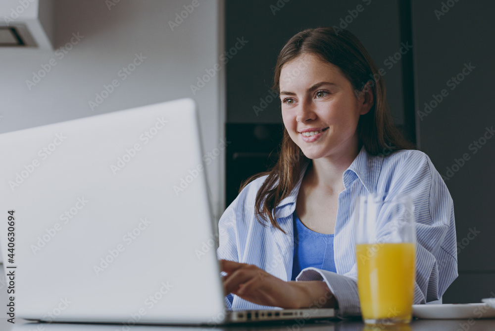 Smiling freelancer copywriter young housewife woman in casual clothes blue shirt use laptop pc computer drink juice look for recipe cook food in light kitchen at home Healthy diet lifestyle concept. Smiling freelancer copywriter young housewife woman in casual clothes blue shirt use laptop pc computer drink juice look for recipe cook food in light kitchen at home Healthy diet lifestyle concept.