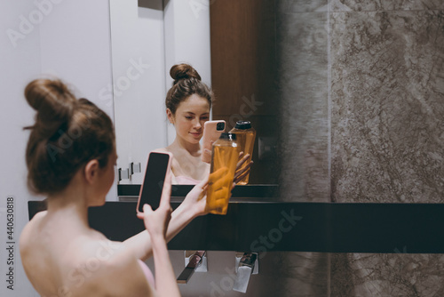 Back rear view half naked topless young woman 20s take photo of shampoo bottle by mobile cell phone reflected in mirror in bathroom do morning routine Skin care healthcare cosmetic procedures concept.