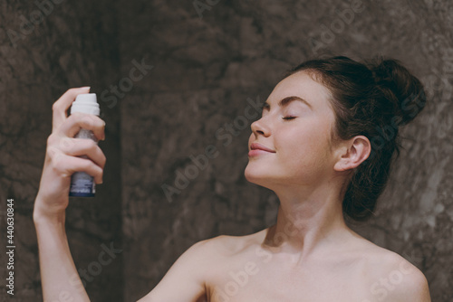 Side view half naked topless young caucasian woman 20s apply on fixative and make-up base on face with closed eye in bathroom brown dark background. Skin care healthcare cosmetic procedures concept