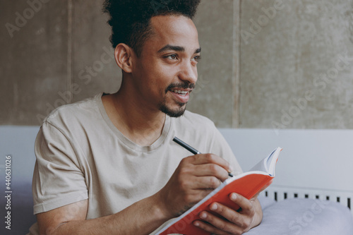 Dreamful creative happy young student african american man 20s in beige t-shirt sitting on sofa indoors apartment write down memories in notebook diary, writing letter, rest on weekends stay at home photo