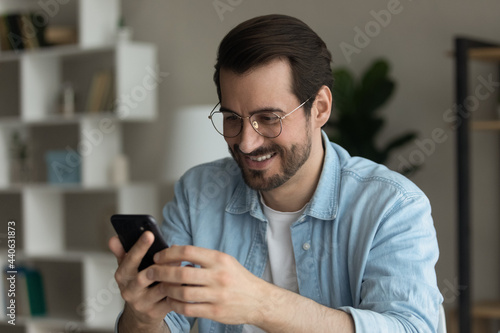 Happy young man in eyewear looking at smartphone screen, feeling excited of reading pleasant media news, enjoying playing mobile game, shopping in internet store or web surfing, modern tech addiction.