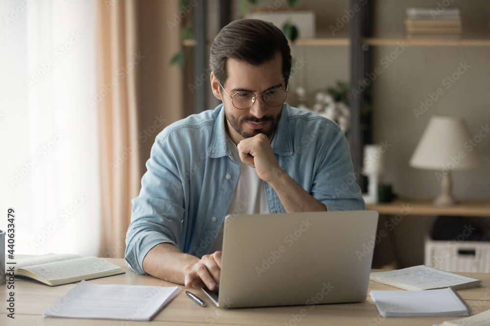 Fototapeta premium Concentrated happy young business man in eyewear looking computer screen, developing online project growth strategy, preparing presentation, web surfing information doing research, working distantly.