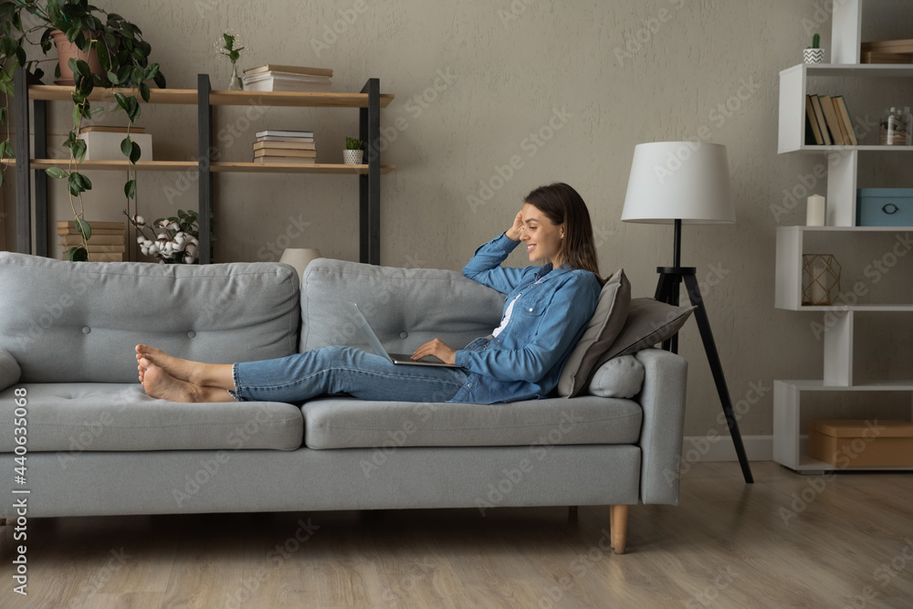 Full length relaxed millennial smiling woman lying on cozy sofa, enjoying using computer software applications, creating new online project, reading media news, playing games, communicating distantly. Full length relaxed millennial smiling woman lying on cozy sofa, enjoying using computer software applications, creating new online project, reading media news, playing games, communicating distantly.