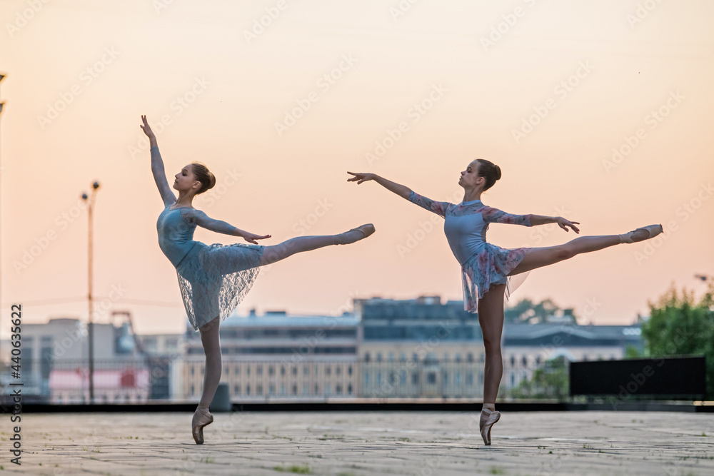 Obraz premium Two young ballerinas dancing in pointe shoes in city against the backdrop of sunset sky