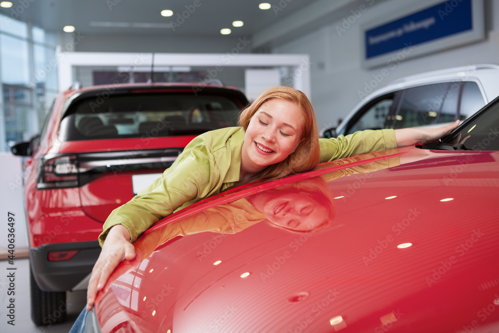Happy young woman looking excited, hugging her new automobile at car dealership