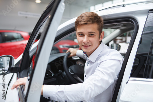 Young man smiling to the camera, getting out of the car at dealership salon