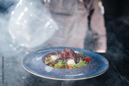 A professional chef serves a freshly prepared salad of tomato and veal greens under a glass hood with thick smoke. Beautiful smoky serving in the restaurant.