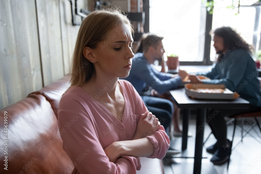 Unhappy abused young caucasian woman sitting on sofa in cafe separately from multiethnic diverse happy people, suffering from bullying, hurt by mockery gossip, unfair attitude or no friends support. Unhappy abused young caucasian woman sitting on sofa in cafe separately from multiethnic diverse happy people, suffering from bullying, hurt by mockery gossip, unfair attitude or no friends support.