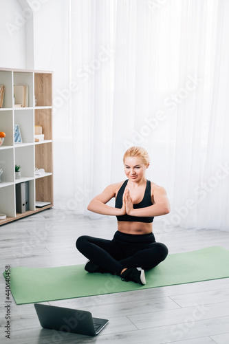 Meditating woman. Home yoga. Online practice. Harmony balance. Healthy body. Happy lady black sportswear sitting lotus pose namaste hands looking laptop in light room interior.