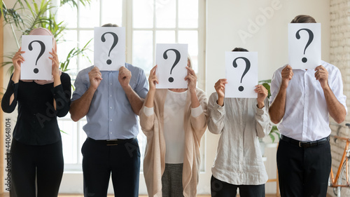 Diverse business people hiding faces behind papers sheets with question marks, standing in row in office, identity and equality at work, candidates waiting for job interview, recruitment concept photo