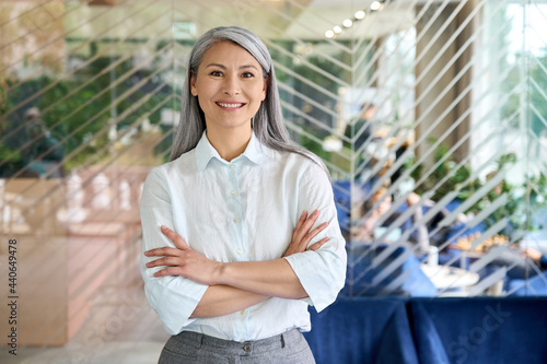 Fotografie Happy smiling confident middle aged Asian older senior female leader businesswoman standing in modern office workplace looking at camera arms crossed