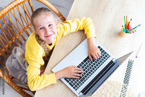 Caucasian beautiful girl 10-11 years old sits in a wicker chair at a desk made of light wood, hands on laptop keyboard, looking at the camera, smiles, top view. A cozy workplace for a student. Online.