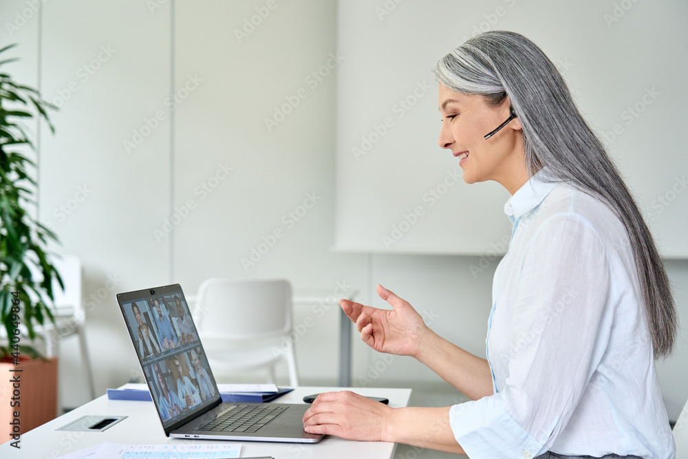 Happy smiling older Asian businesswoman ceo executive manager in headset on corporate videoconference with multicultural colleagues using laptop in modern corporation office. Business career concept. Happy smiling older Asian businesswoman ceo executive manager in headset on corporate videoconference with multicultural colleagues using laptop in modern corporation office. Business career concept.