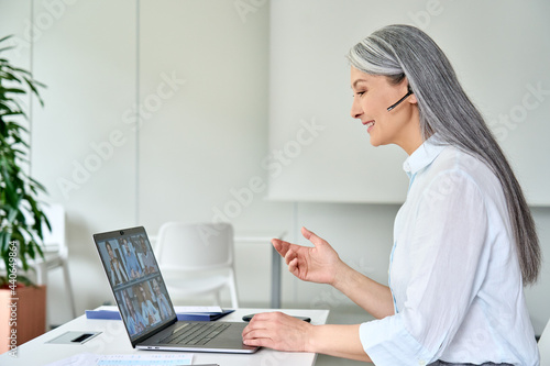 Happy smiling older Asian businesswoman ceo executive manager in headset on corporate videoconference with multicultural colleagues using laptop in modern corporation office. Business career concept.