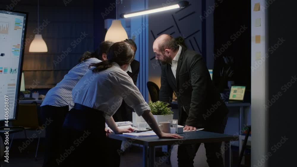 Diverse business teamwork standing at conference table checking management graphs paperwork late at night. Multi ethnic coworkers planning company presentation in meeting room brainstorming ideas Diverse business teamwork standing at conference table checking management graphs paperwork late at night. Multi ethnic coworkers planning company presentation in meeting room brainstorming ideas