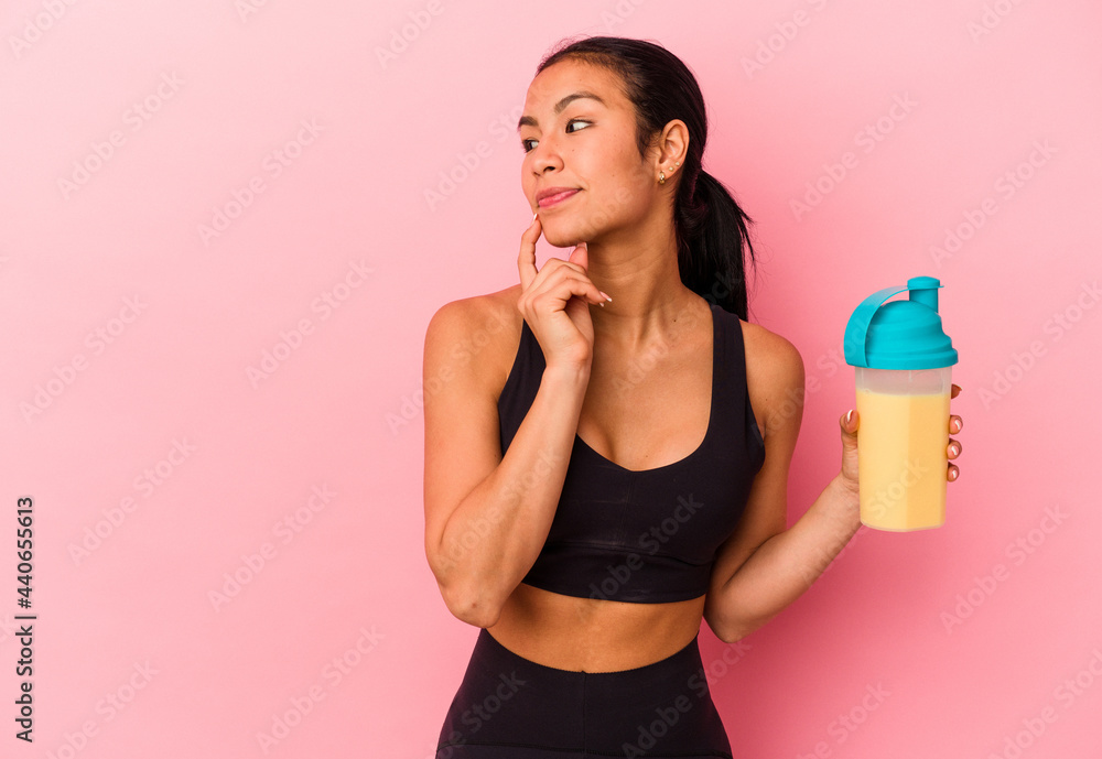 Young Venezuelan woman drinking a protein shake isolated on pink background relaxed thinking about something looking at a copy space. Young Venezuelan woman drinking a protein shake isolated on pink background relaxed thinking about something looking at a copy space.