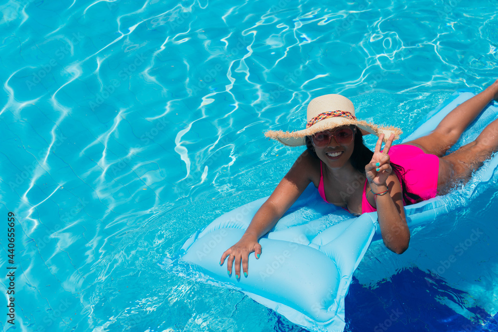 Smiling young woman in bikini, wearing straw hat relaxing on inflatable in pool. Attractive woman in swimwear stands in the sun on tropical vacation. Woman sunbathing at a luxury resort.