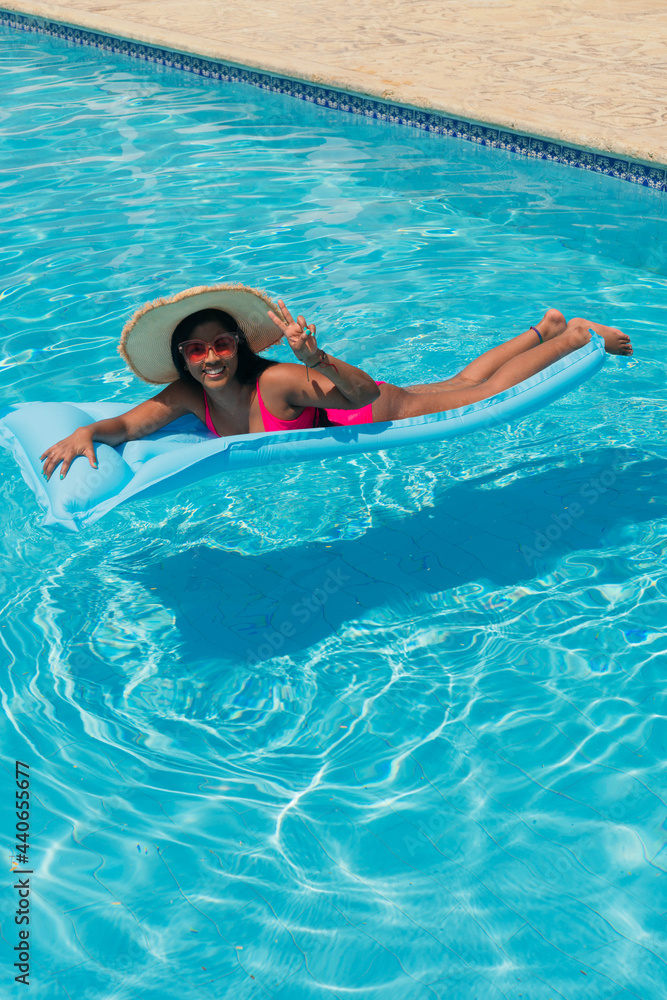 Young smiling African American girl in bikini, wearing straw hat relaxing on inflatable in swimming pool.