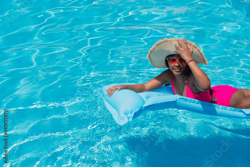 Young smiling African girl in bikini, wearing straw hat relaxing on inflatable in swimming pool.
