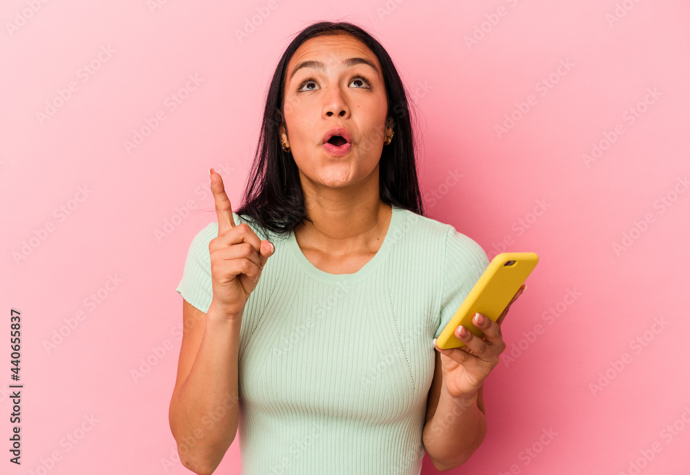 Young Venezuelan woman holding a mobile phone isolated on pink background pointing upside with opened mouth. Young Venezuelan woman holding a mobile phone isolated on pink background pointing upside with opened mouth.
