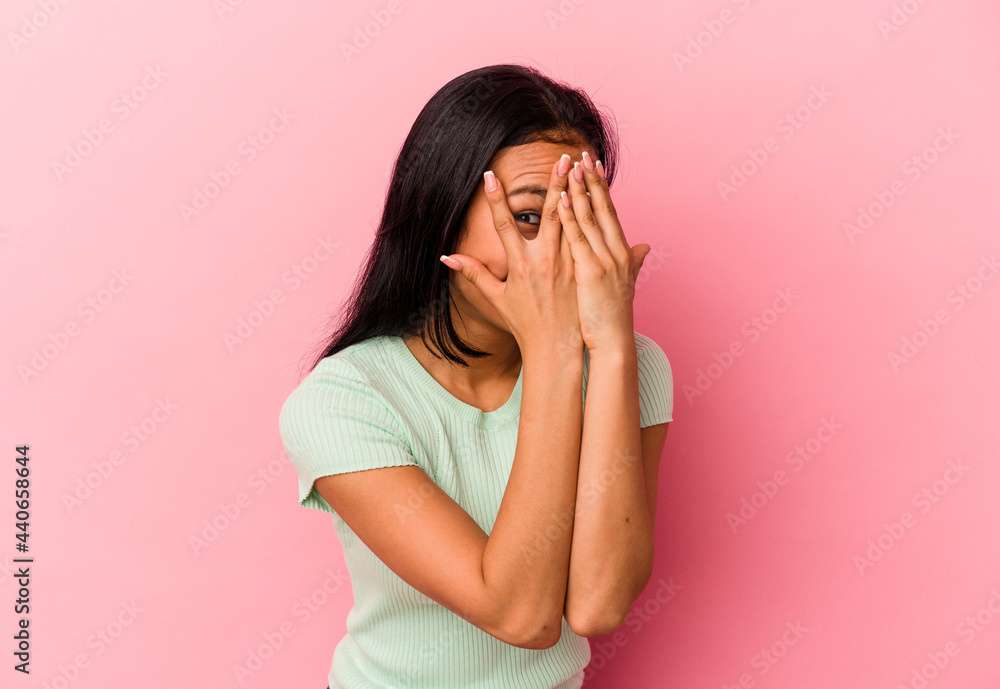 Young Venezuelan woman isolated on pink background blink through fingers frightened and nervous.