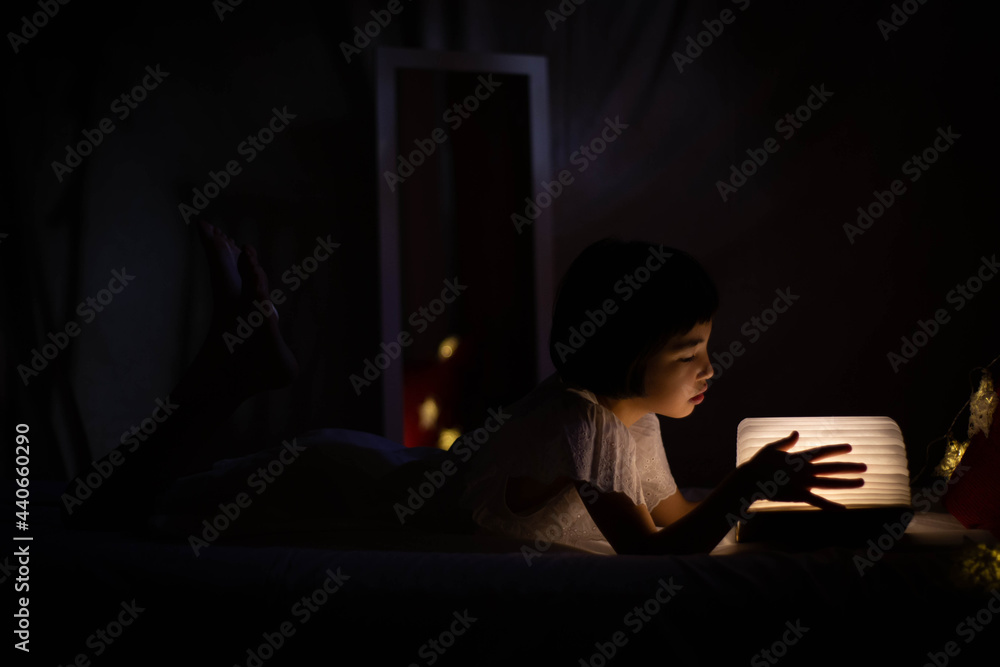 Bella niña asiática leyendo libro luminoso en la cama